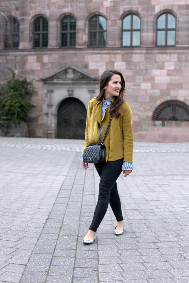 Ein fröhlicher Frühlingslook mit senfgelber Trenchcoatjacke, gestreifter Trompetenbluse und silbernen spitzen Ballerinas