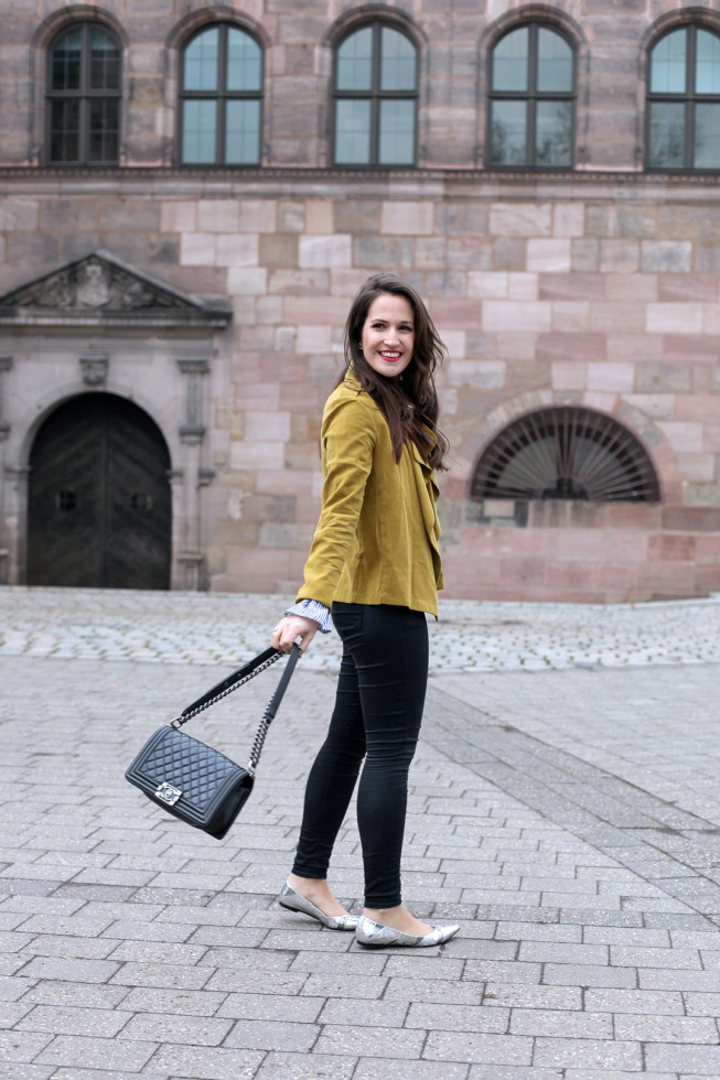 Ein fröhlicher Frühlingslook mit senfgelber Trenchcoatjacke, gestreifter Trompetenbluse und silbernen spitzen Ballerinas