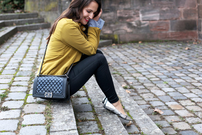 Ein fröhlicher Frühlingslook mit senfgelber Trenchcoatjacke, gestreifter Trompetenbluse und silbernen spitzen Ballerinas