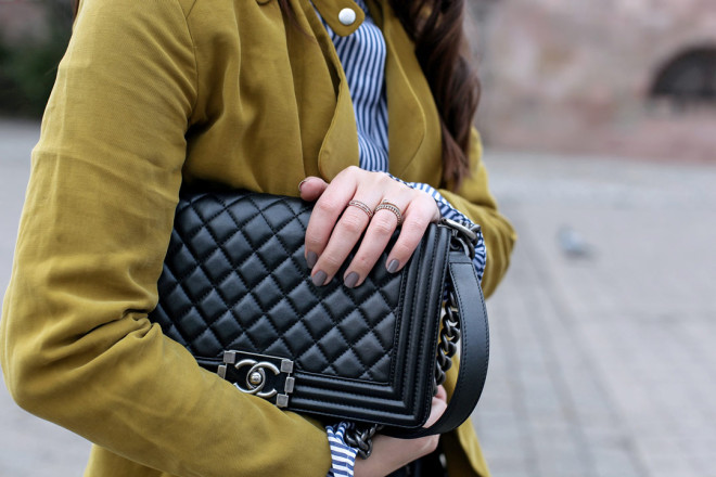 Ein fröhlicher Frühlingslook mit senfgelber Trenchcoatjacke, gestreifter Trompetenbluse und silbernen spitzen Ballerinas