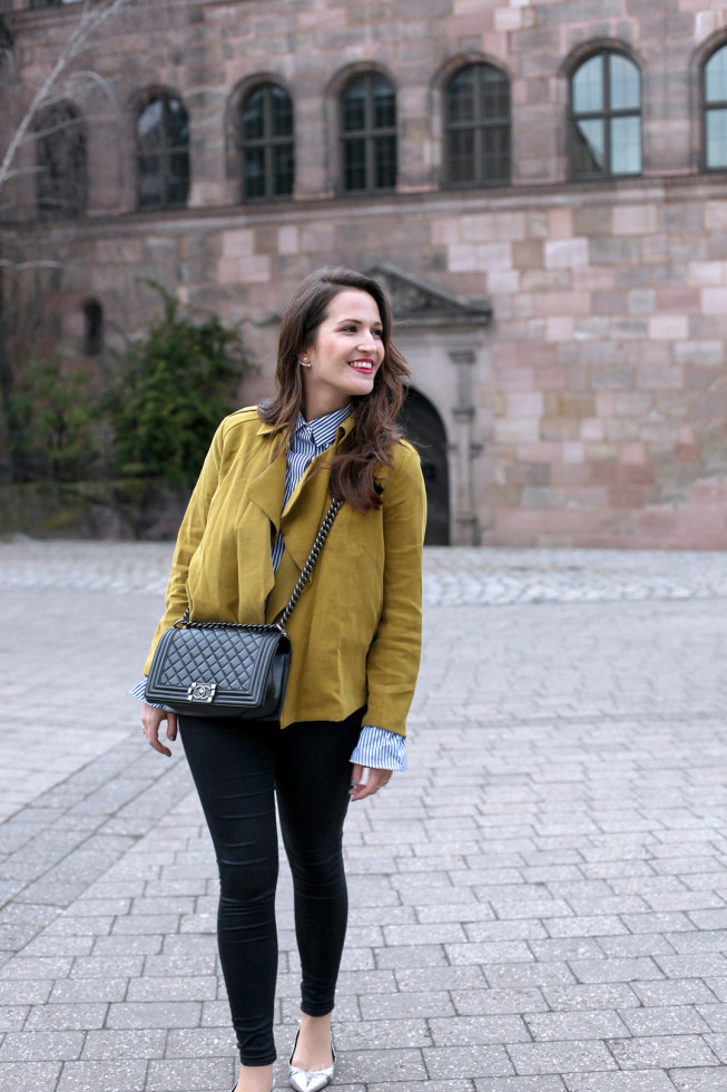 Ein fröhlicher Frühlingslook mit senfgelber Trenchcoatjacke, gestreifter Trompetenbluse und silbernen spitzen Ballerinas